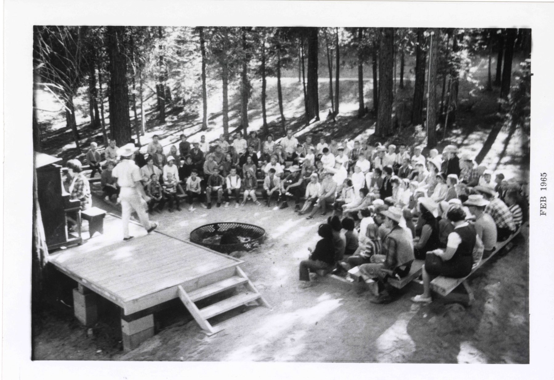A 1965 black-and-white photograph from above: a few hundred campers on wooden benches in a half-circle around a fire ring, a speaker on a plank stage and a woman at an upright piano, under tall pines.