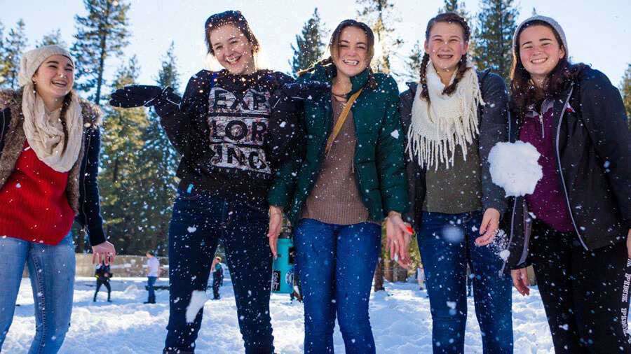A row of teenagers in winter coats throwing snow in the air at camp.